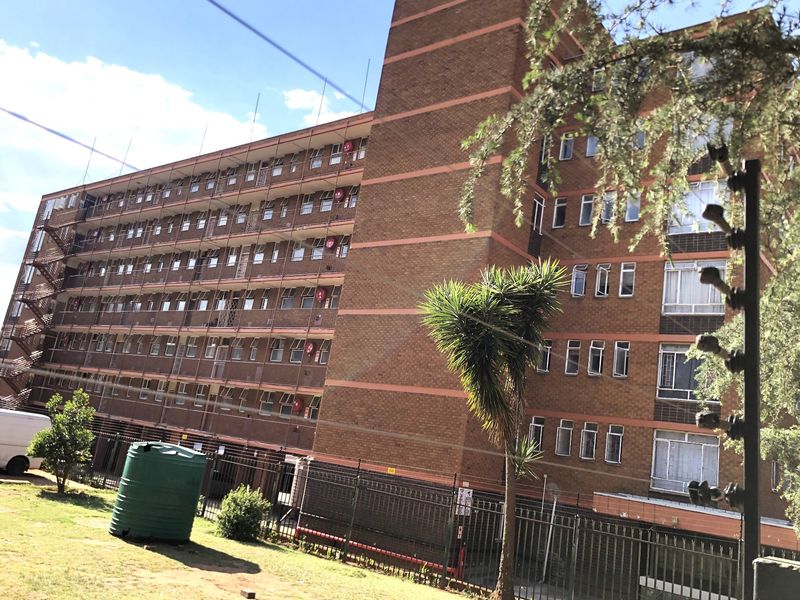 Exterior view of a multi-story brick apartment building featuring a metal fire escape, security fencing, and a water storage tank on the premises.