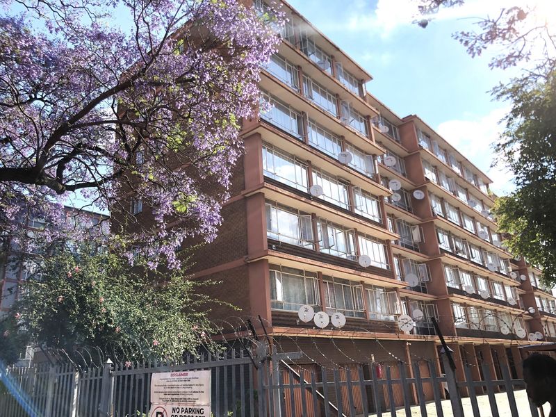 Exterior view of a multi-story apartment building featuring brick facade, large metal-framed windows, and perimeter security fencing with razor wire.