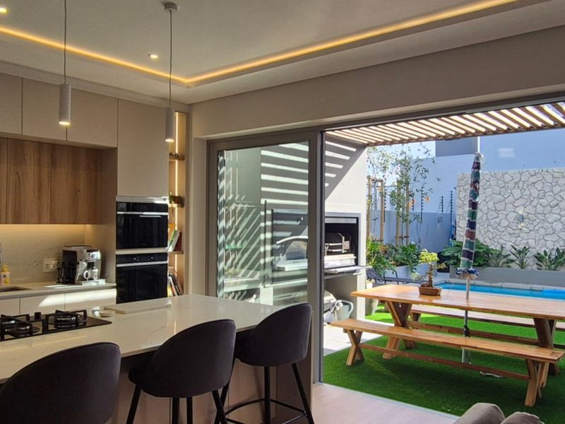 Modern open-plan kitchen featuring a central island and high-end appliances, seamlessly flowing onto a covered patio with a built-in braai and pool area.