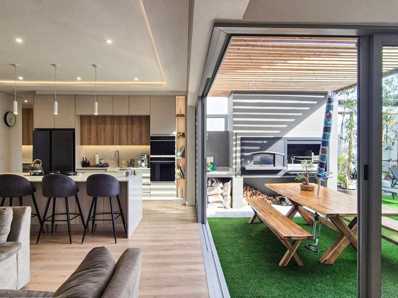 Modern open-plan living area featuring a sleek kitchen with island, flowing seamlessly through large sliding doors to a covered patio with built-in braai.