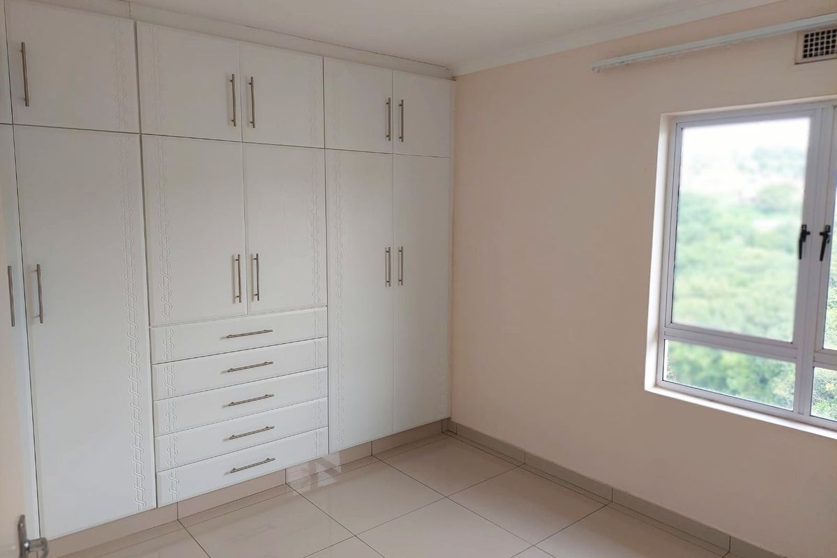 A bright and empty bedroom featuring modern white built-in cupboards, neutral tiled flooring, and a large window providing natural light.