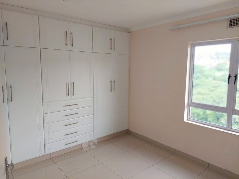 A bright and empty bedroom featuring modern white built-in cupboards, neutral tiled flooring, and a large window providing natural light.