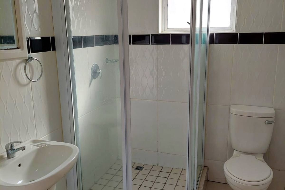 A functional bathroom featuring a pedestal basin, a glass-enclosed shower, and a standard toilet, finished with neutral wall and floor tiles.