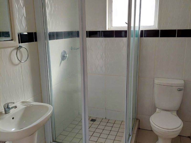 A functional bathroom featuring a pedestal basin, a glass-enclosed shower, and a standard toilet, finished with neutral wall and floor tiles.