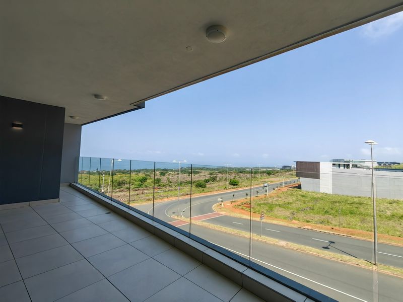Spacious covered balcony featuring modern tiled flooring and a frameless glass balustrade, offering expansive views of the surrounding landscape and coastal area.
