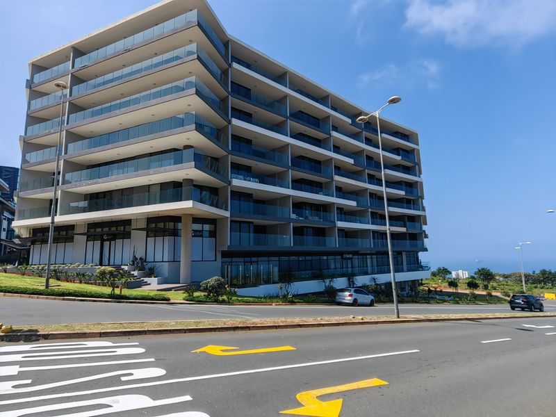 Modern multi-story apartment building featuring extensive glass balcony balustrades and a contemporary architectural design, situated along a paved street.