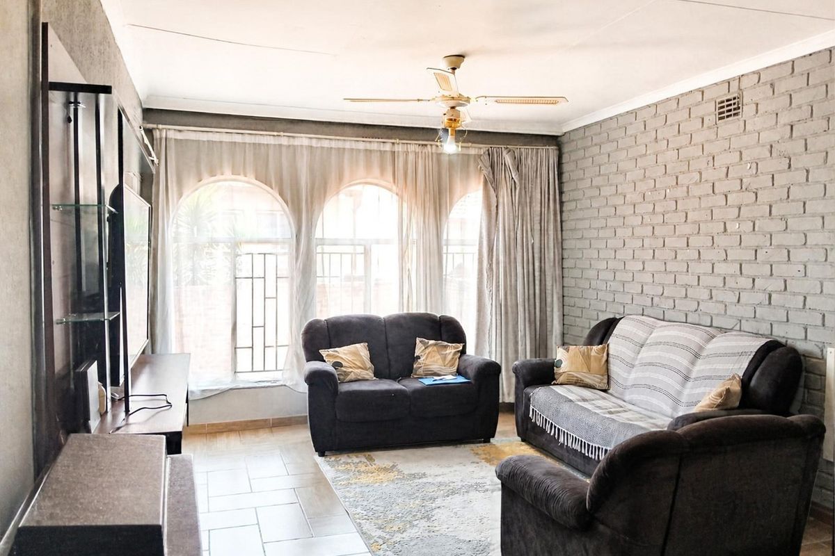 A living room featuring an exposed brick wall, tiled flooring, a ceiling fan, and large arched windows with security bars.