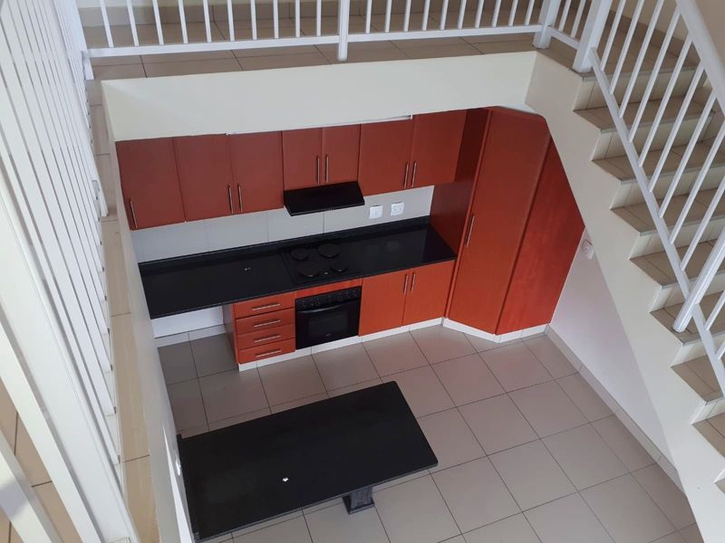 A compact kitchen featuring wooden cabinetry, black granite countertops, built-in oven and hob, situated beneath a staircase in an open-plan apartment layout.