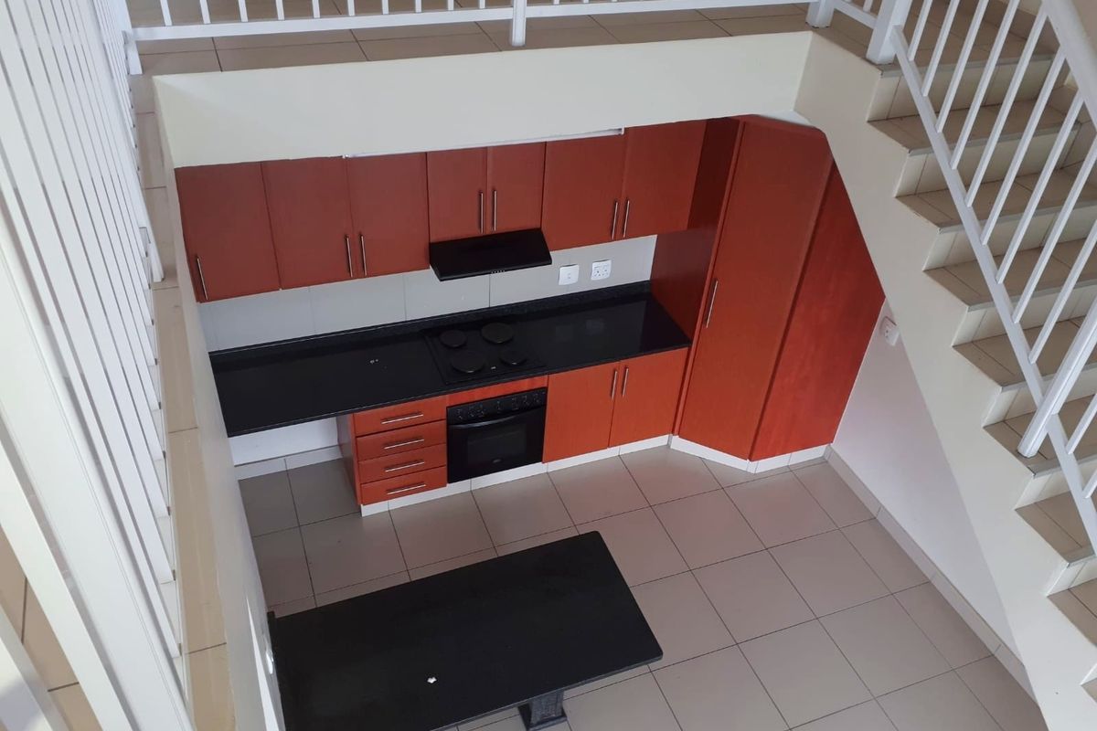 A compact kitchen featuring wooden cabinetry, black granite countertops, built-in oven and hob, situated beneath a staircase in an open-plan apartment layout.