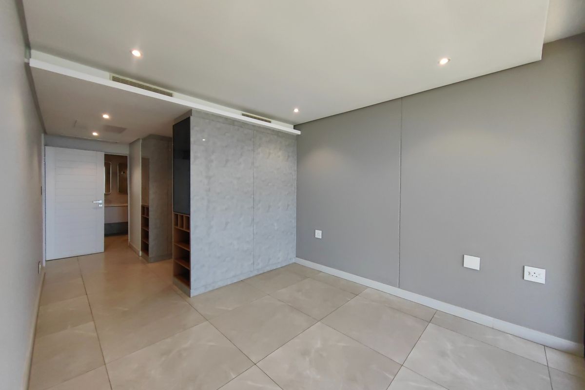 A modern, spacious bedroom featuring neutral grey walls, large format floor tiles, recessed lighting, and built-in cabinetry with integrated shelving.