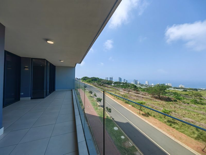 Spacious covered balcony featuring modern tiled flooring, a sleek glass balustrade, and expansive views of the surrounding landscape and coastline.