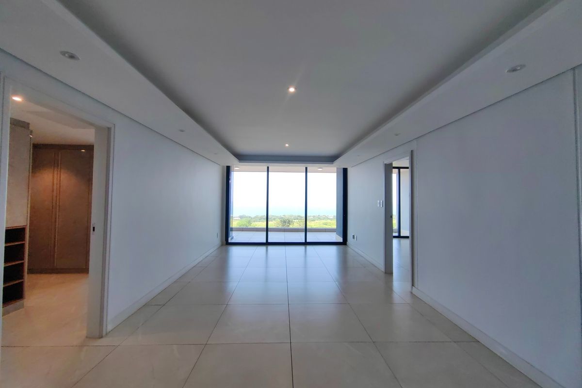 A bright, spacious, and empty living area featuring large format floor tiles, recessed ceiling lighting, and expansive sliding glass doors leading to a balcony.