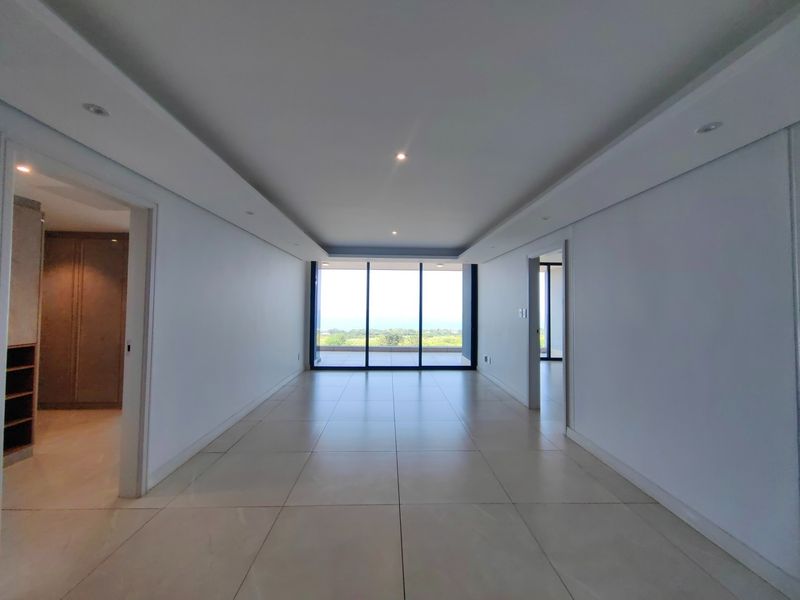 A bright, spacious, and empty living area featuring large format floor tiles, recessed ceiling lighting, and expansive sliding glass doors leading to a balcony.
