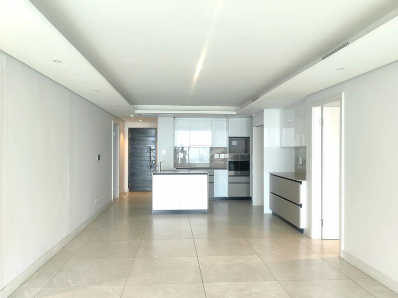 A modern, open-plan living and kitchen area featuring sleek white cabinetry, a central kitchen island, built-in appliances, and neutral tiled flooring throughout.