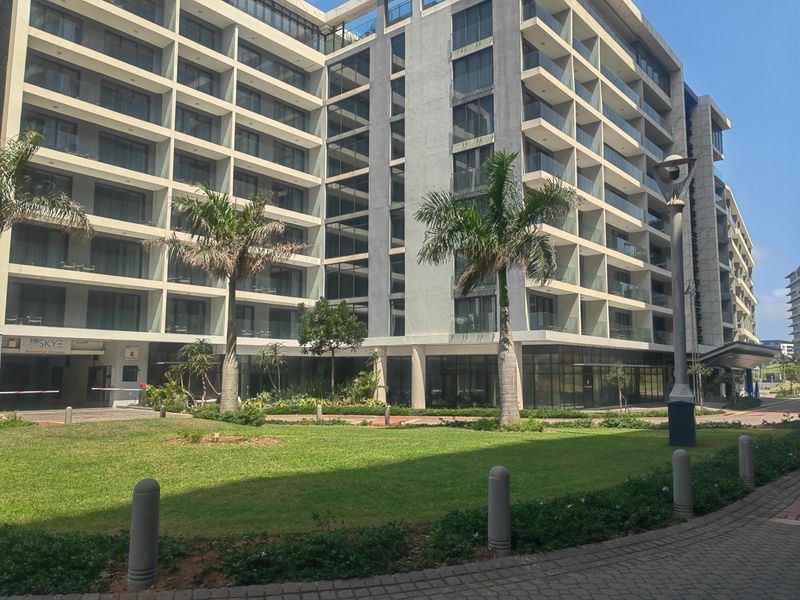 A modern multi-story apartment building featuring glass balconies, a well-maintained landscaped garden, and paved pedestrian walkways.