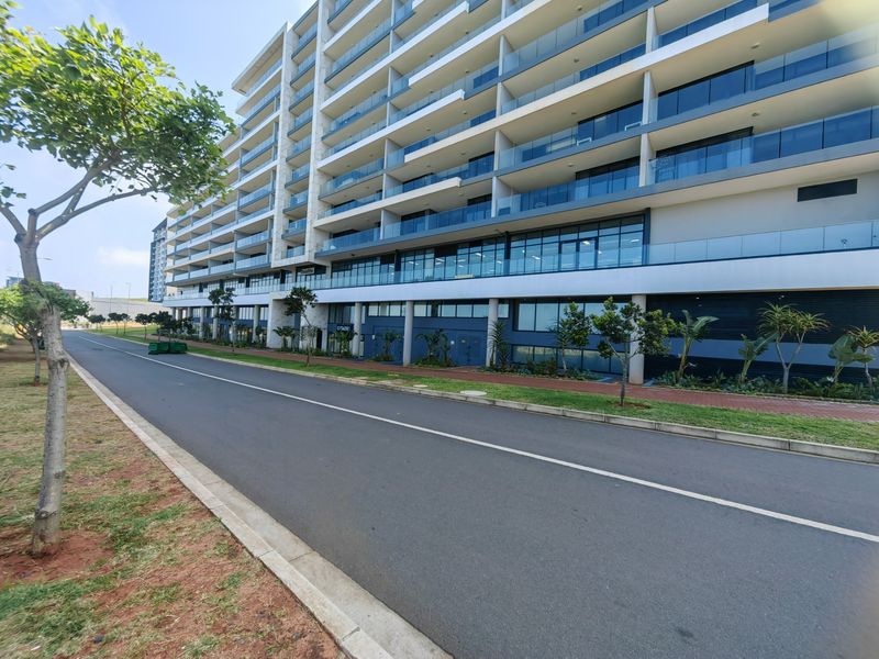 A modern multi-story apartment building featuring glass balconies and a clean, contemporary facade, situated along a paved street with landscaped surroundings.