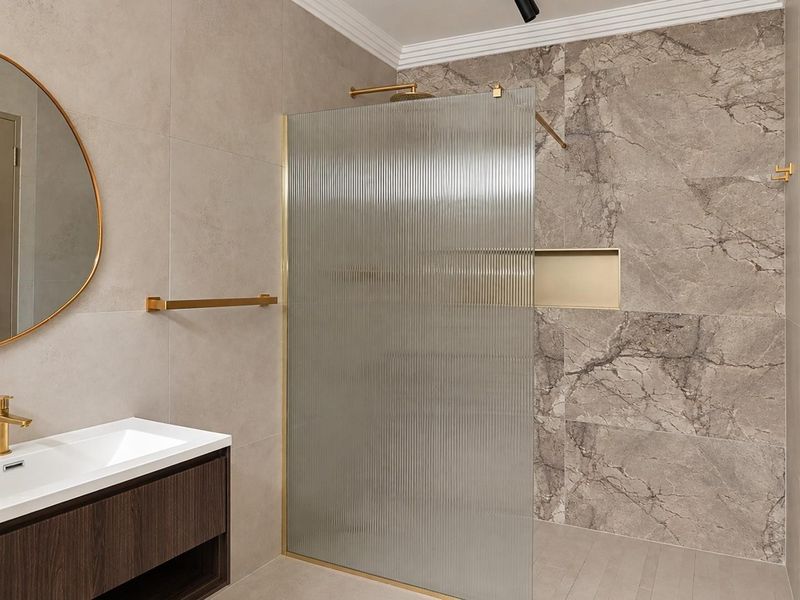 Modern bathroom featuring a walk-in shower with fluted glass screen, brass fixtures, built-in niche, and a sleek wall-mounted vanity with integrated basin.