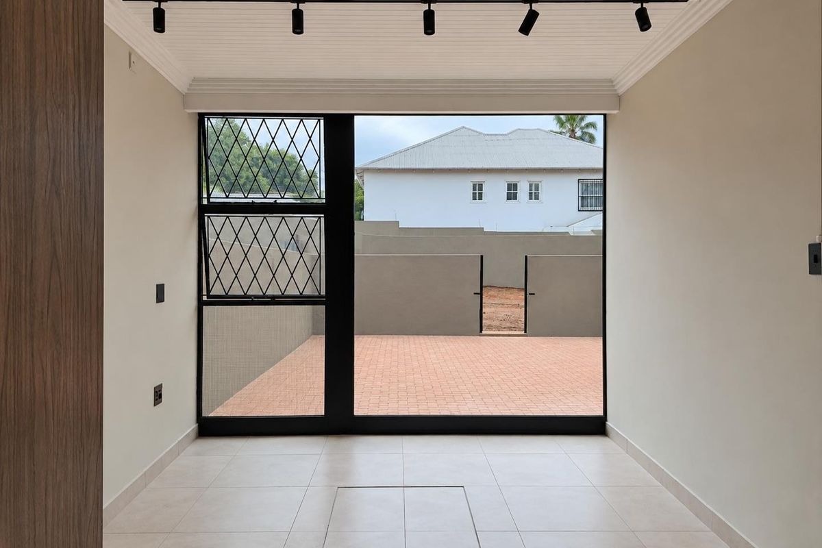 A bright, empty living area featuring modern track lighting, neutral tiled floors, and a large glass door opening onto a paved outdoor space.