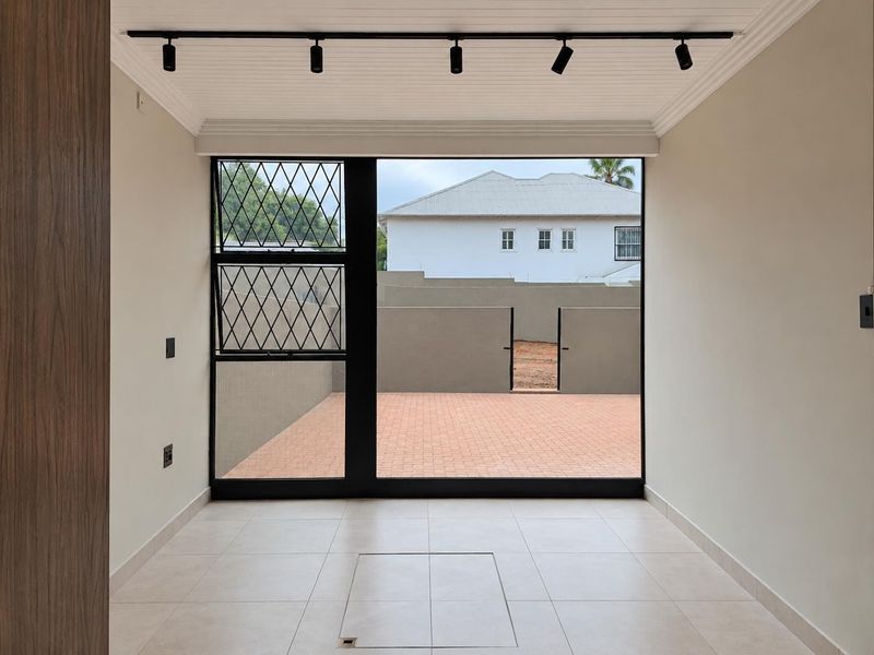 A bright, empty living area featuring modern track lighting, neutral tiled floors, and a large glass door opening onto a paved outdoor space.