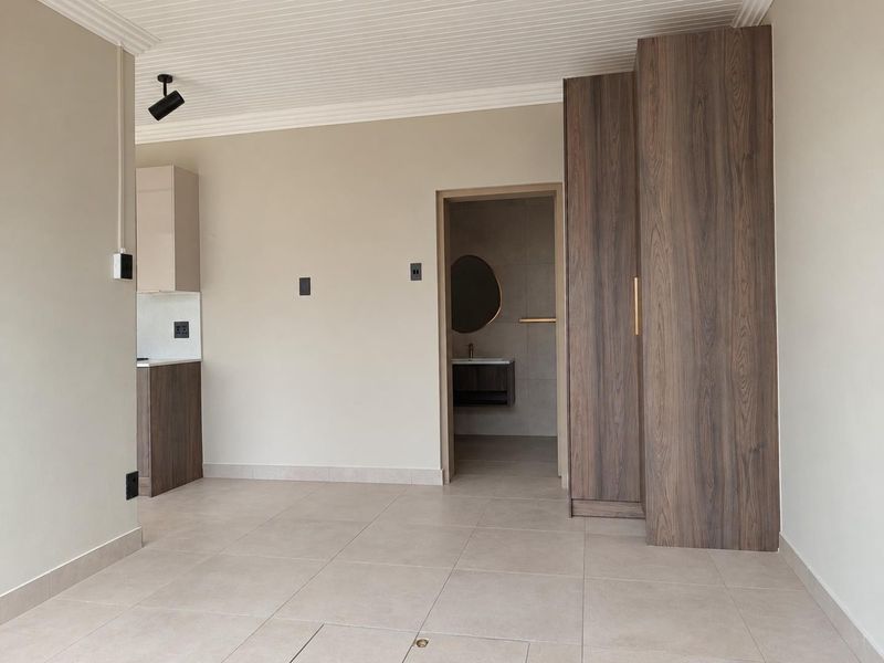 Modern open-plan living area featuring neutral tiled flooring, built-in wood-grain cabinetry, and clean finishes, with direct access to an adjoining bathroom.