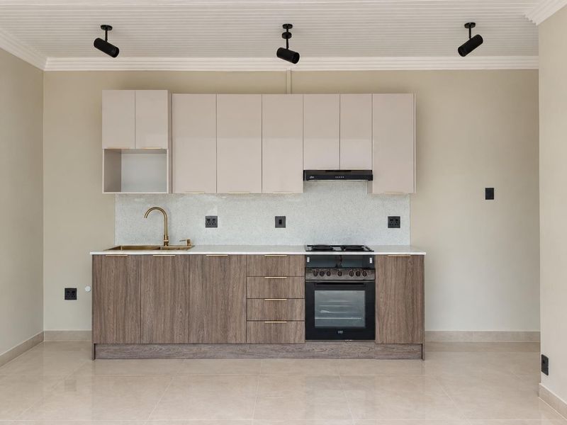 Modern kitchen featuring two-tone cabinetry, stone countertops, integrated oven, gas hob, and brass fixtures, set against a neutral backdrop with track lighting.