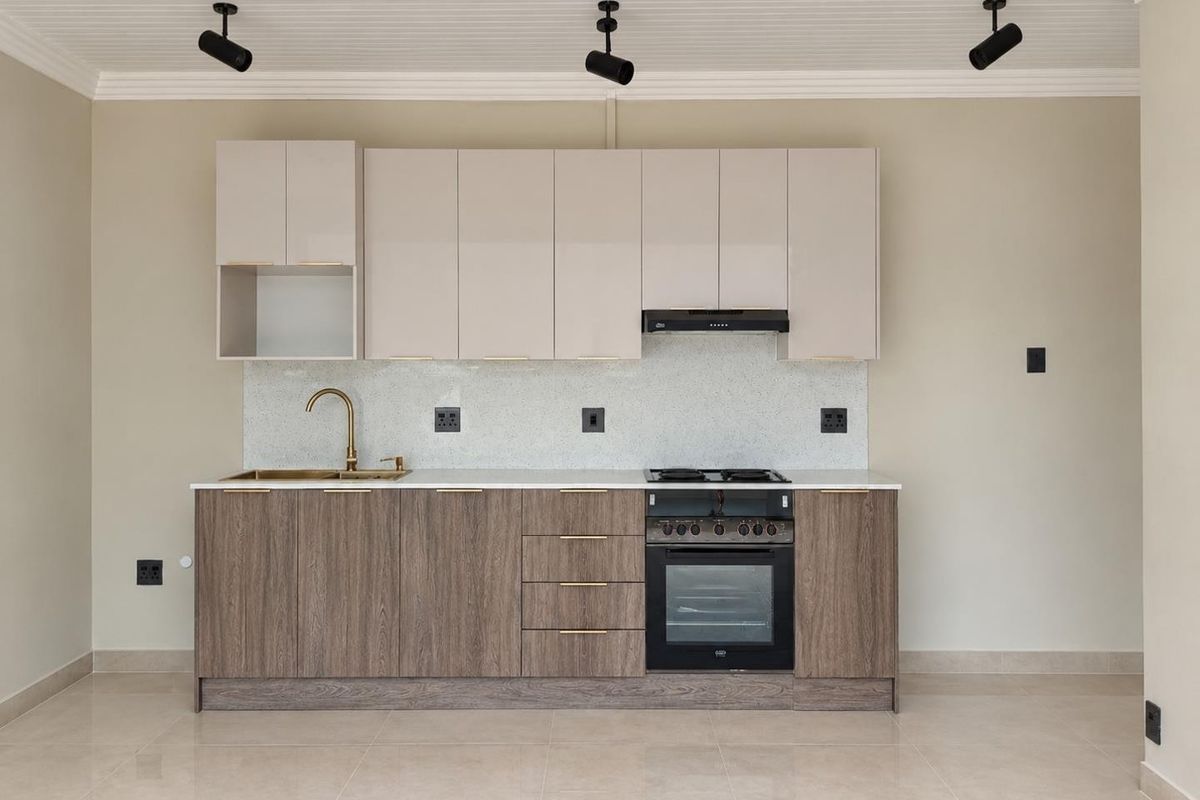 Modern kitchen featuring two-tone cabinetry, stone countertops, integrated oven, gas hob, and brass fixtures, set against a neutral backdrop with track lighting.
