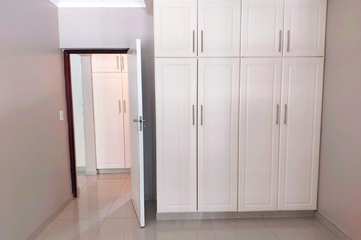 A clean, empty bedroom featuring neutral tiled flooring and ample built-in cupboard storage, with a view into an adjacent hallway.