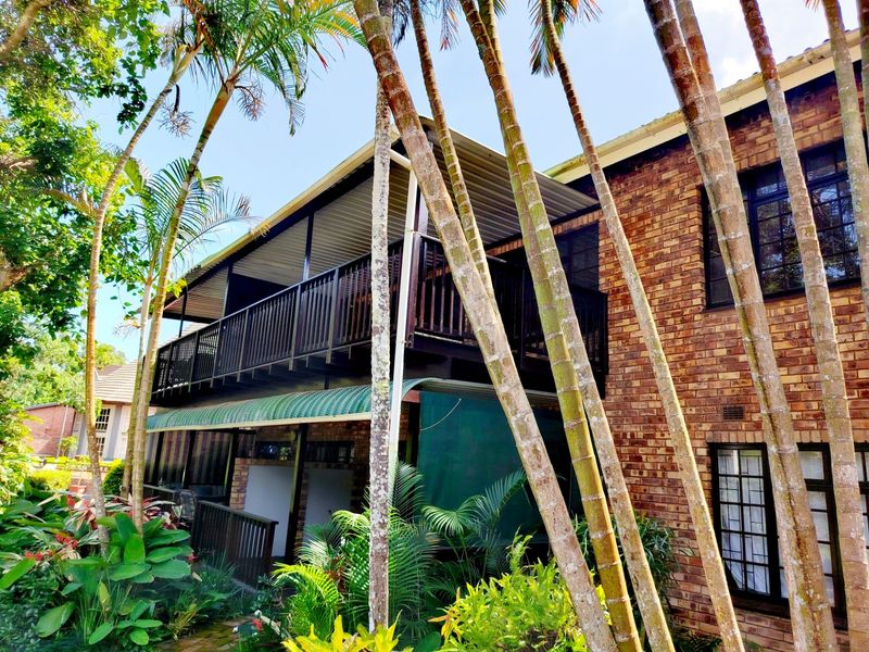Rear view of a face brick apartment building featuring a covered balcony with timber railings and established garden surroundings.