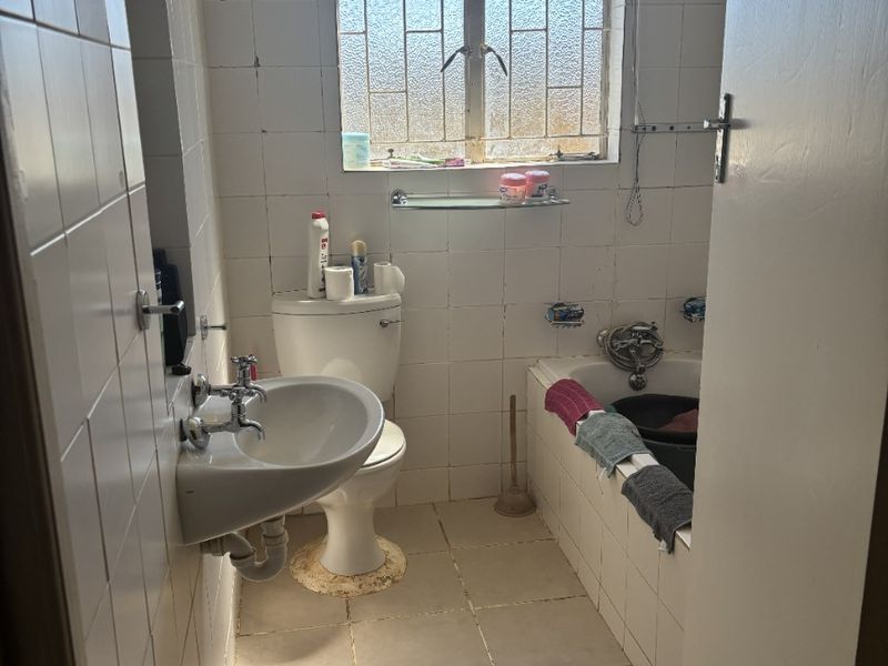 A compact bathroom featuring a wall-mounted basin, toilet, and built-in bathtub with tiled finishes, requiring cosmetic updates to improve its overall condition.