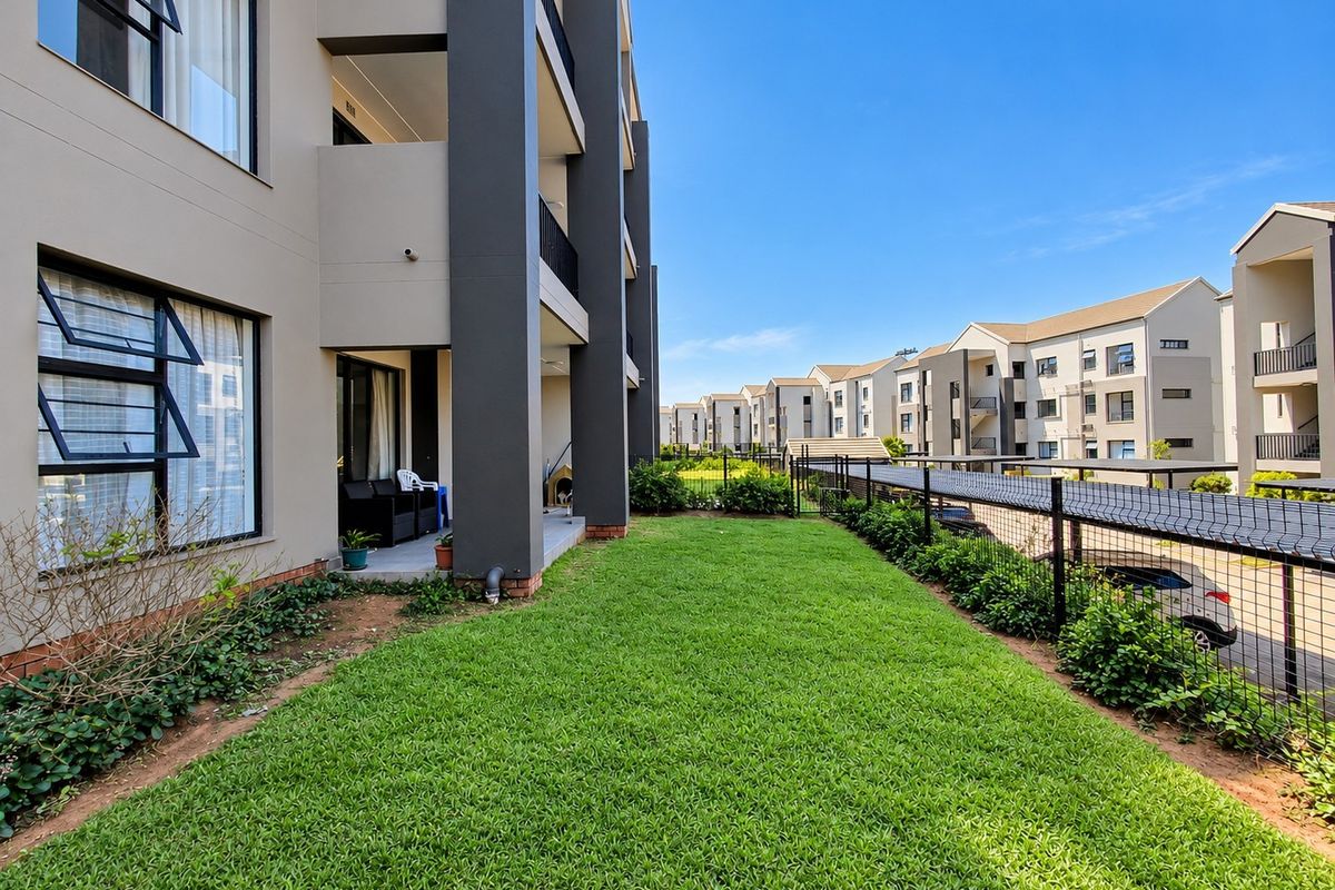 A view of the rear exterior of the apartment building, featuring a private lawn area, covered patio, and secure perimeter fencing.