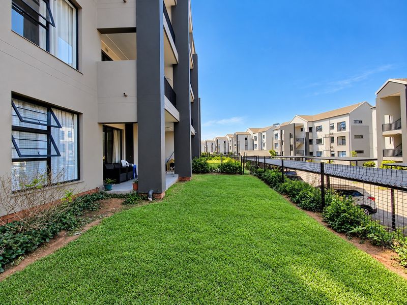 A view of the rear exterior of the apartment building, featuring a private lawn area, covered patio, and secure perimeter fencing.