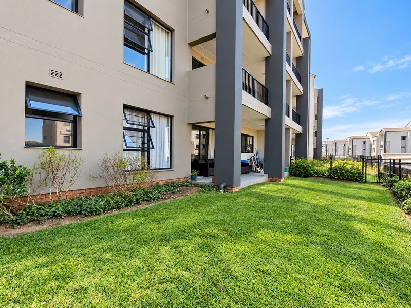 Rear view of a modern apartment building featuring a private ground-floor patio, metal balcony railings, and a well-maintained communal lawn area.