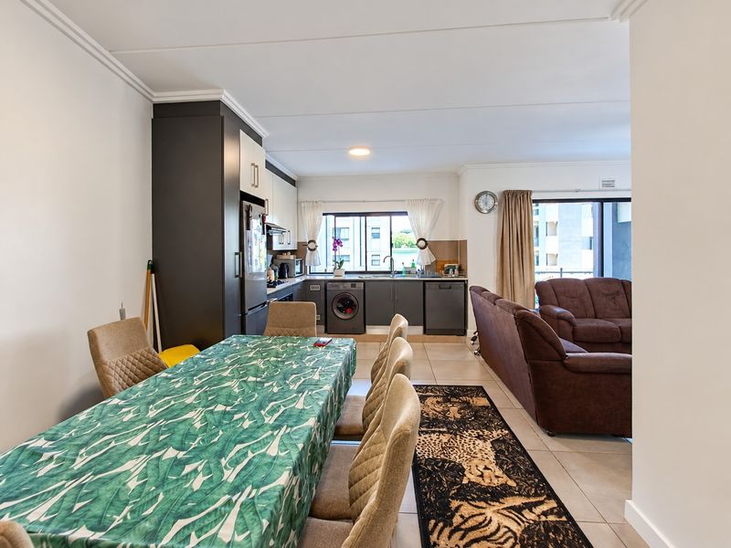 An open-plan living and dining area featuring a modern kitchen with built-in cabinetry, integrated appliances, and tiled flooring, leading to a balcony.