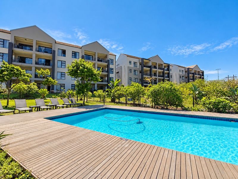 A well-maintained communal swimming pool area featuring timber decking, landscaped gardens, and views of the apartment complex buildings.