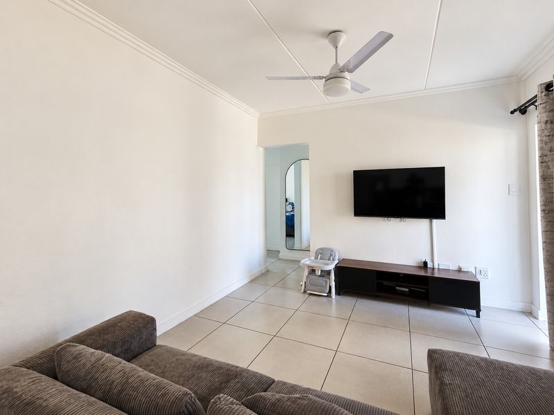 A bright living room featuring neutral tiled flooring, a ceiling fan, and coved ceiling details, providing a clean and functional space.