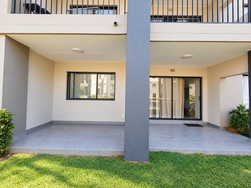 A covered ground-floor patio featuring tiled flooring, a sliding glass door entrance, and a metal balustrade overlooking a small garden area.