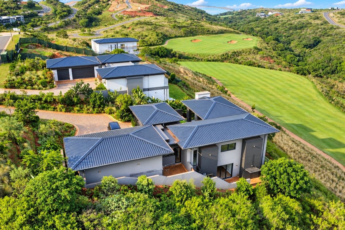 Aerial view of a modern residential property featuring a metal roof, paved driveway, double garage, and landscaped garden, situated adjacent to a golf course.
