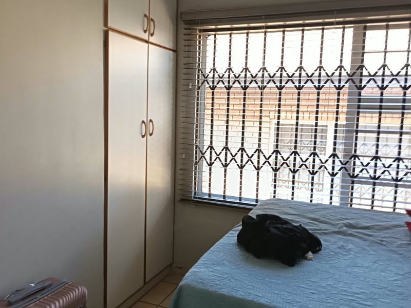 A functional bedroom featuring built-in cupboards, tiled flooring, and a window fitted with security bars and venetian blinds.