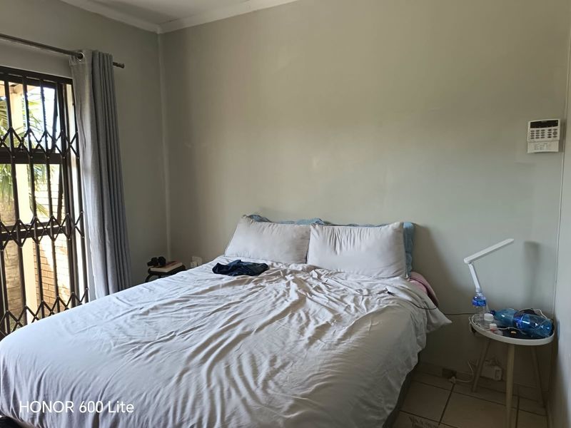 A functional bedroom featuring tiled flooring, a security gate on the window, and a wall-mounted alarm system keypad.