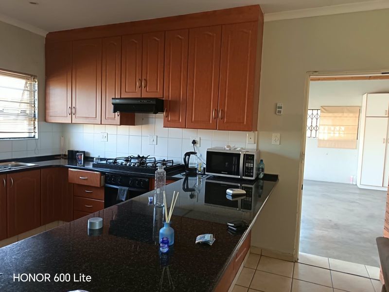 A functional kitchen featuring wooden cabinetry, dark granite countertops, a gas hob with extractor fan, and tiled flooring, leading into an adjacent living area.