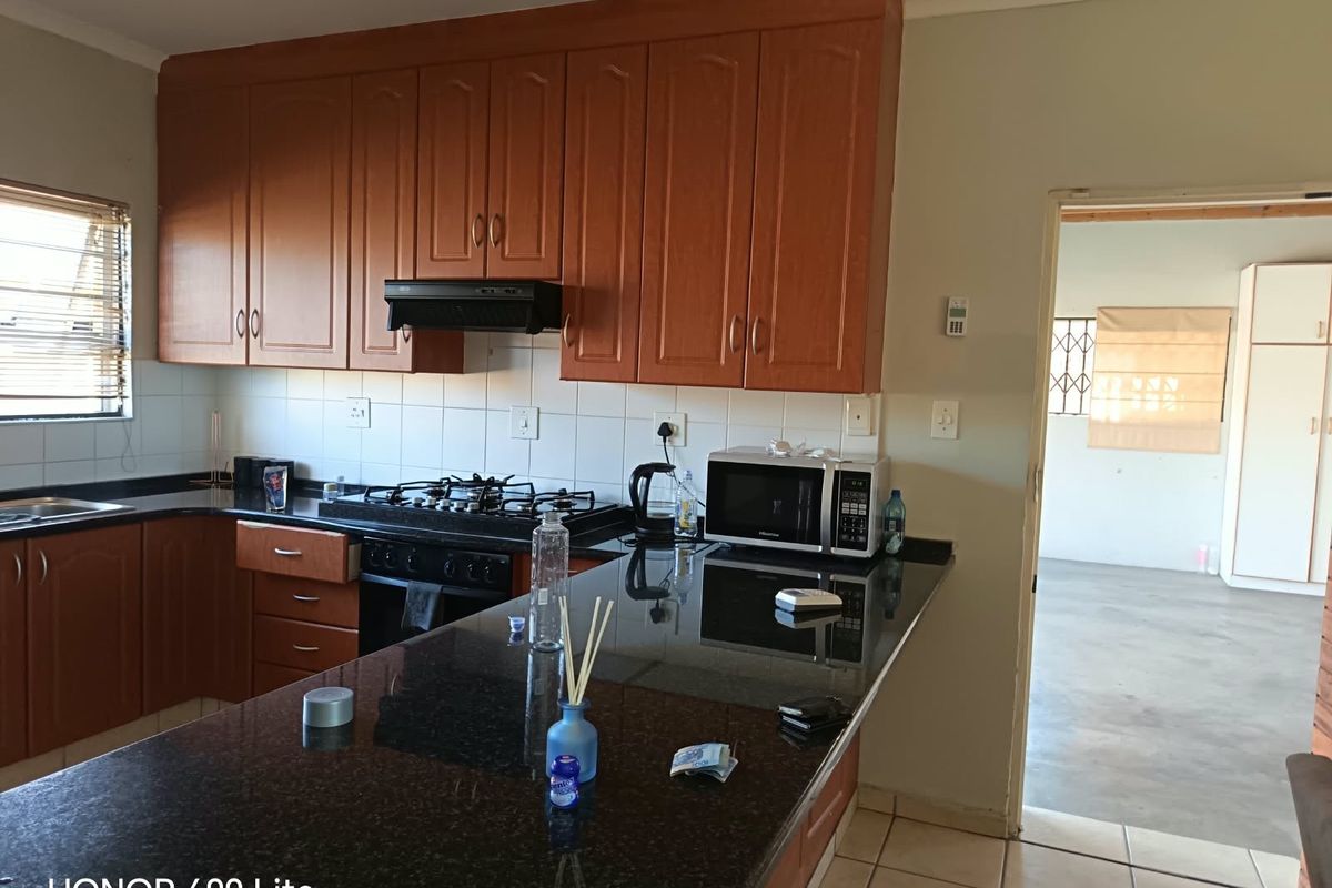 A functional kitchen featuring wooden cabinetry, dark granite countertops, a gas hob with extractor fan, and tiled flooring, leading into an adjacent living area.