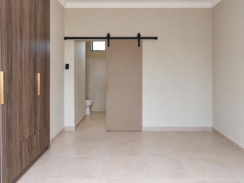 A modern, minimalist bedroom featuring neutral tiled floors, a large built-in wardrobe, and a contemporary sliding barn door leading to an en-suite bathroom.