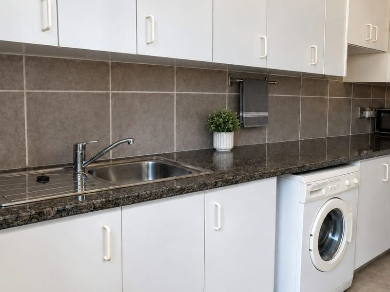 A functional kitchen featuring white cabinetry, granite countertops, a stainless steel sink, and an integrated washing machine with neutral tiled finishes.