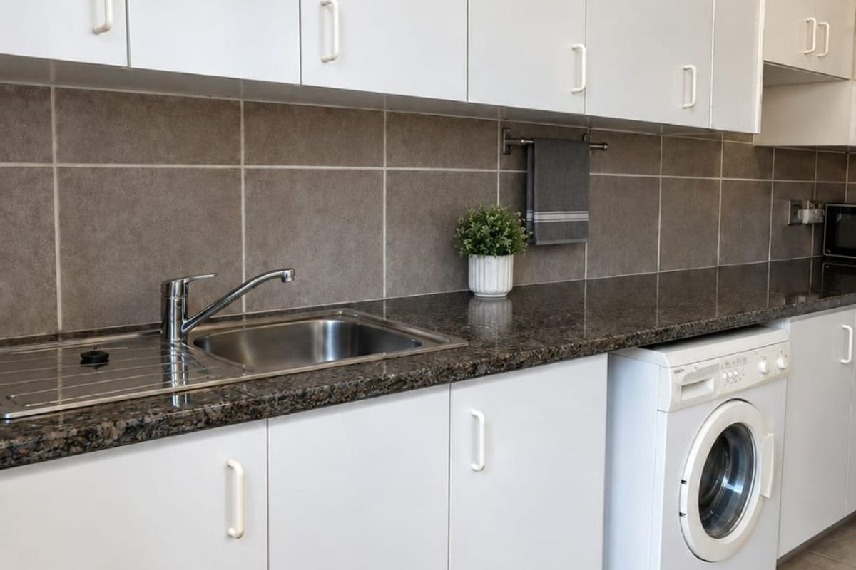 A functional kitchen featuring white cabinetry, granite countertops, a stainless steel sink, and an integrated washing machine with neutral tiled finishes.