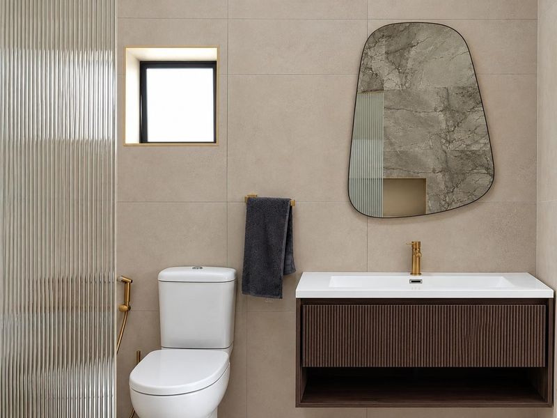 Modern bathroom featuring a wall-mounted vanity with integrated basin, brass fixtures, fluted glass shower screen, and neutral large-format wall tiles.