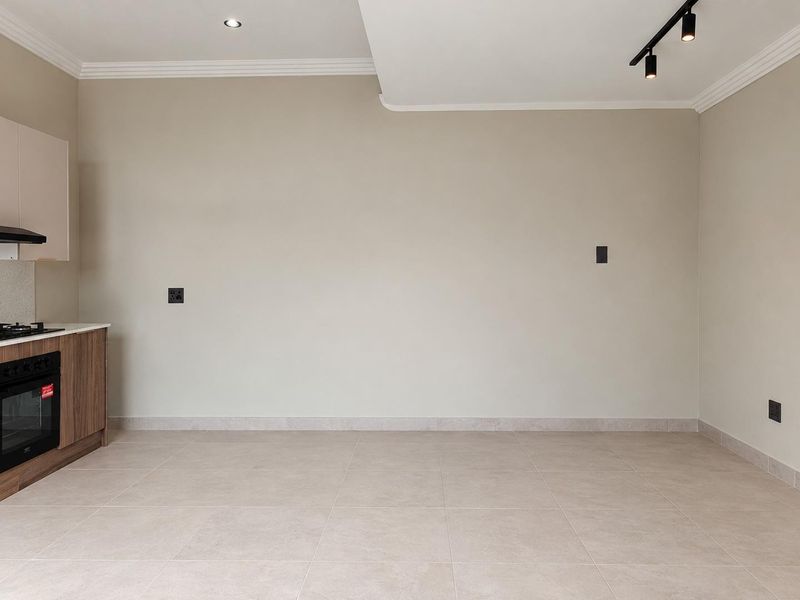 A bright, modern open-plan living area featuring neutral tiled floors, contemporary track lighting, and a compact, well-appointed kitchen section with built-in appliances.