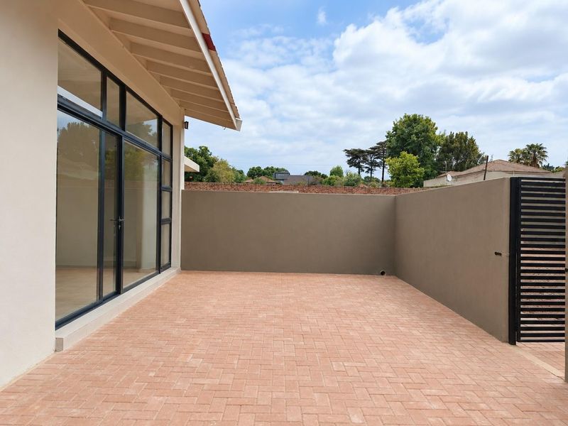 A clean, paved outdoor patio area featuring a boundary wall, metal gate, and large sliding glass doors providing access to the interior.