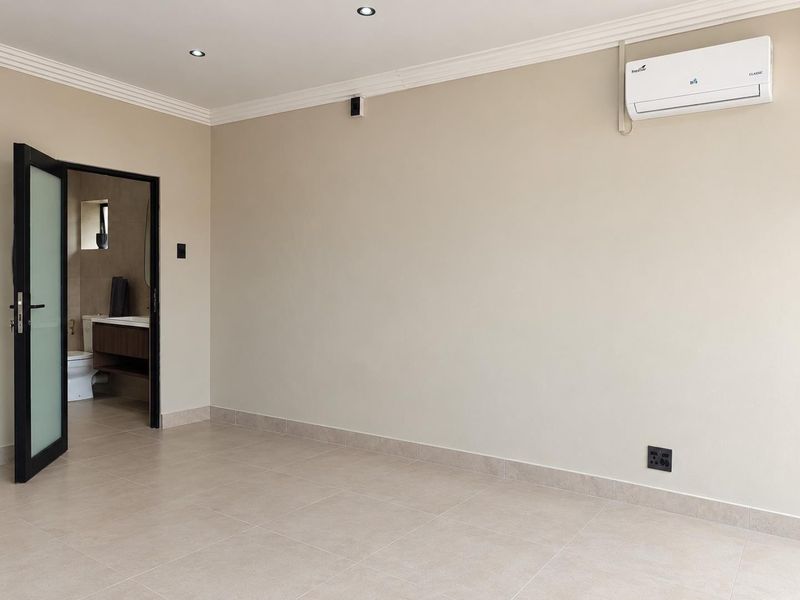 A clean, neutral bedroom featuring tiled floors, recessed lighting, and a wall-mounted air conditioning unit, with an adjoining bathroom visible through a frosted glass door.
