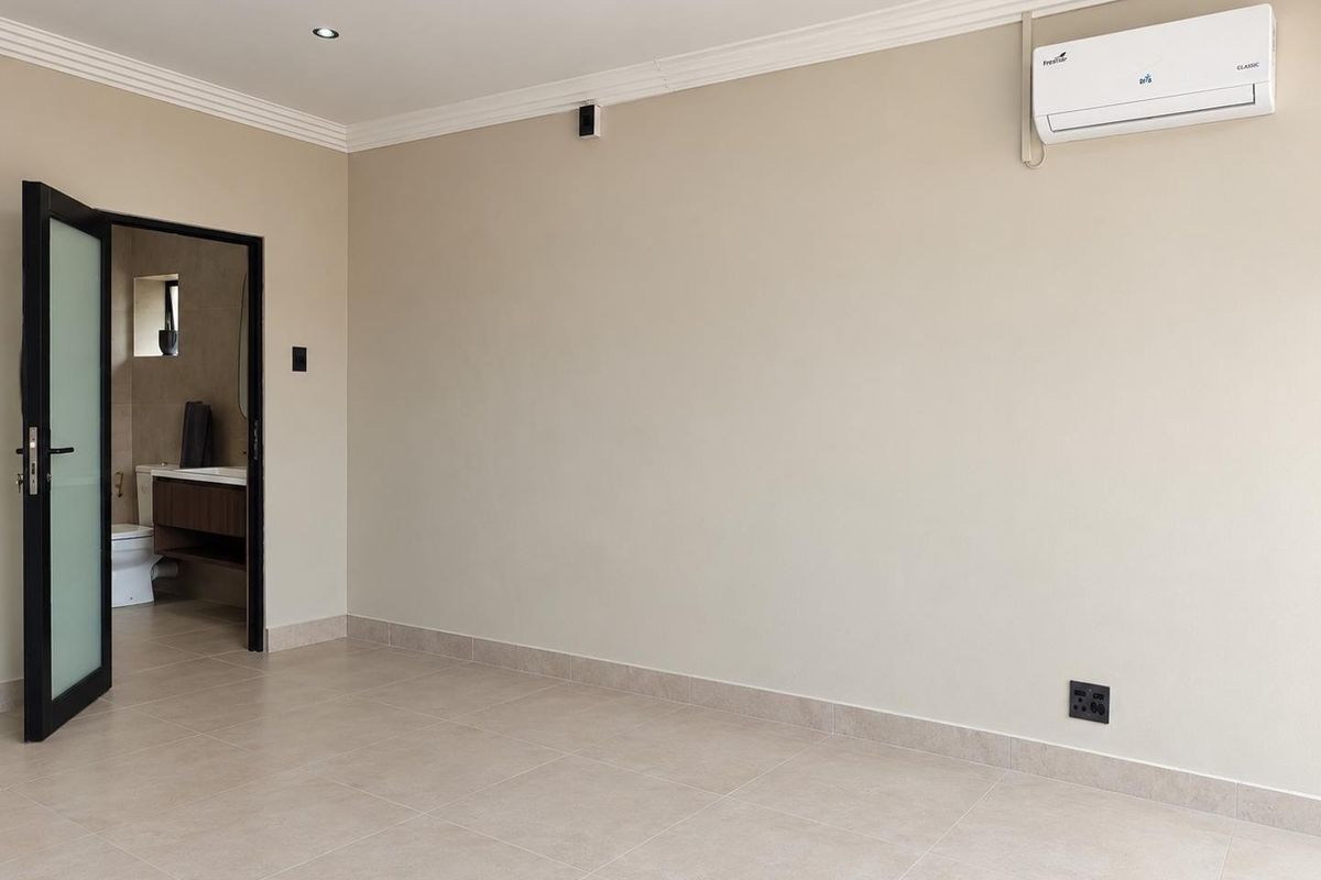 A clean, neutral bedroom featuring tiled floors, recessed lighting, and a wall-mounted air conditioning unit, with an adjoining bathroom visible through a frosted glass door.