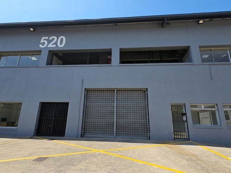 Front elevation of an industrial unit featuring a large roller shutter door, security gates, and concrete driveway access.